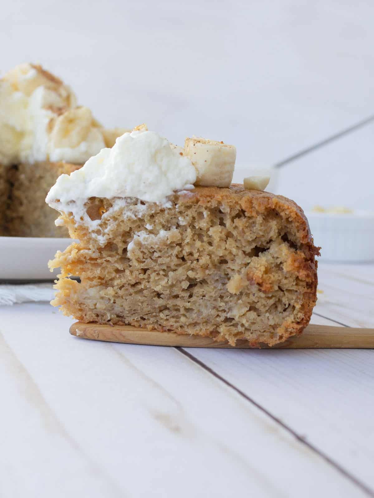 Slice of banana cake on a wood spatula, with whipped cream and bananas on top.