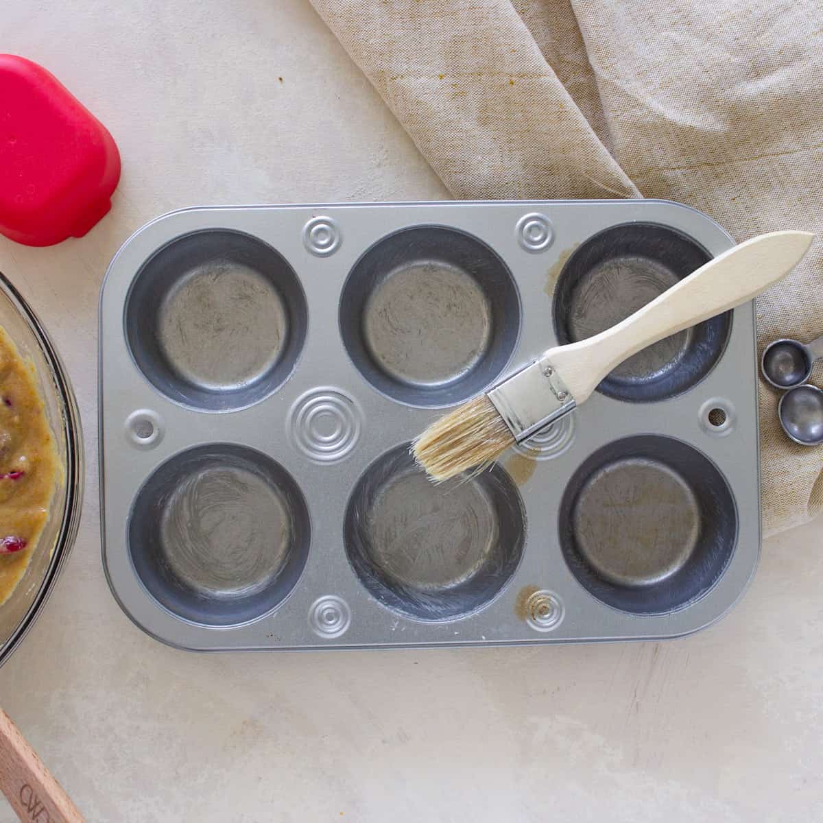 A six cell muffin tin that has been brushed with butter and a pastry brush sitting on top.