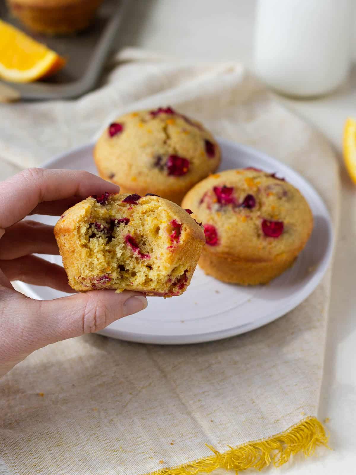 A hand holding a muffins with a bite taken out of it and two muffins on a plate in the background.