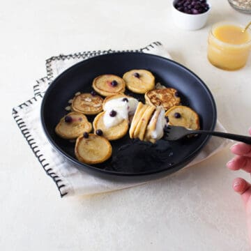 A plate of mini oatmeal pancakes on a black plate topped with yogurt and blueberries and a forkful of pancakes.