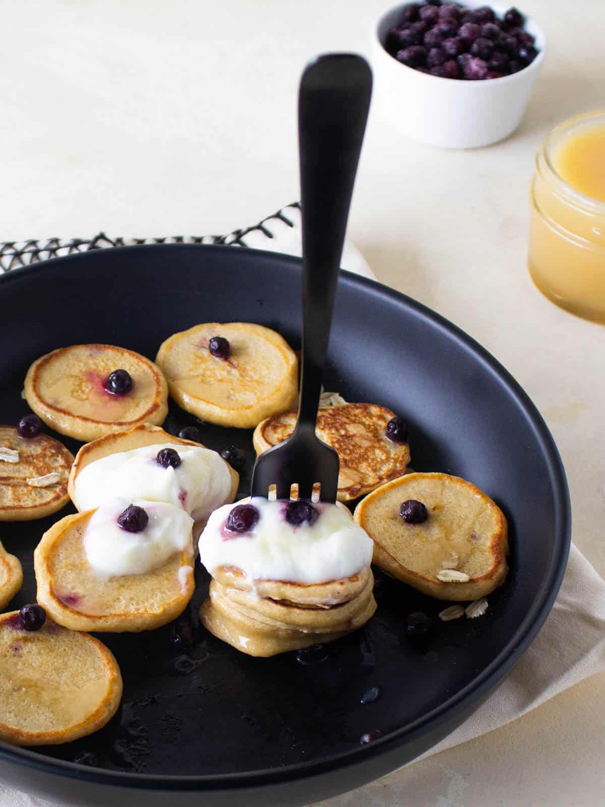 A plate of mini oatmeal pancakes on a black plate topped with yogurt and blueberries and a forkful of pancakes.