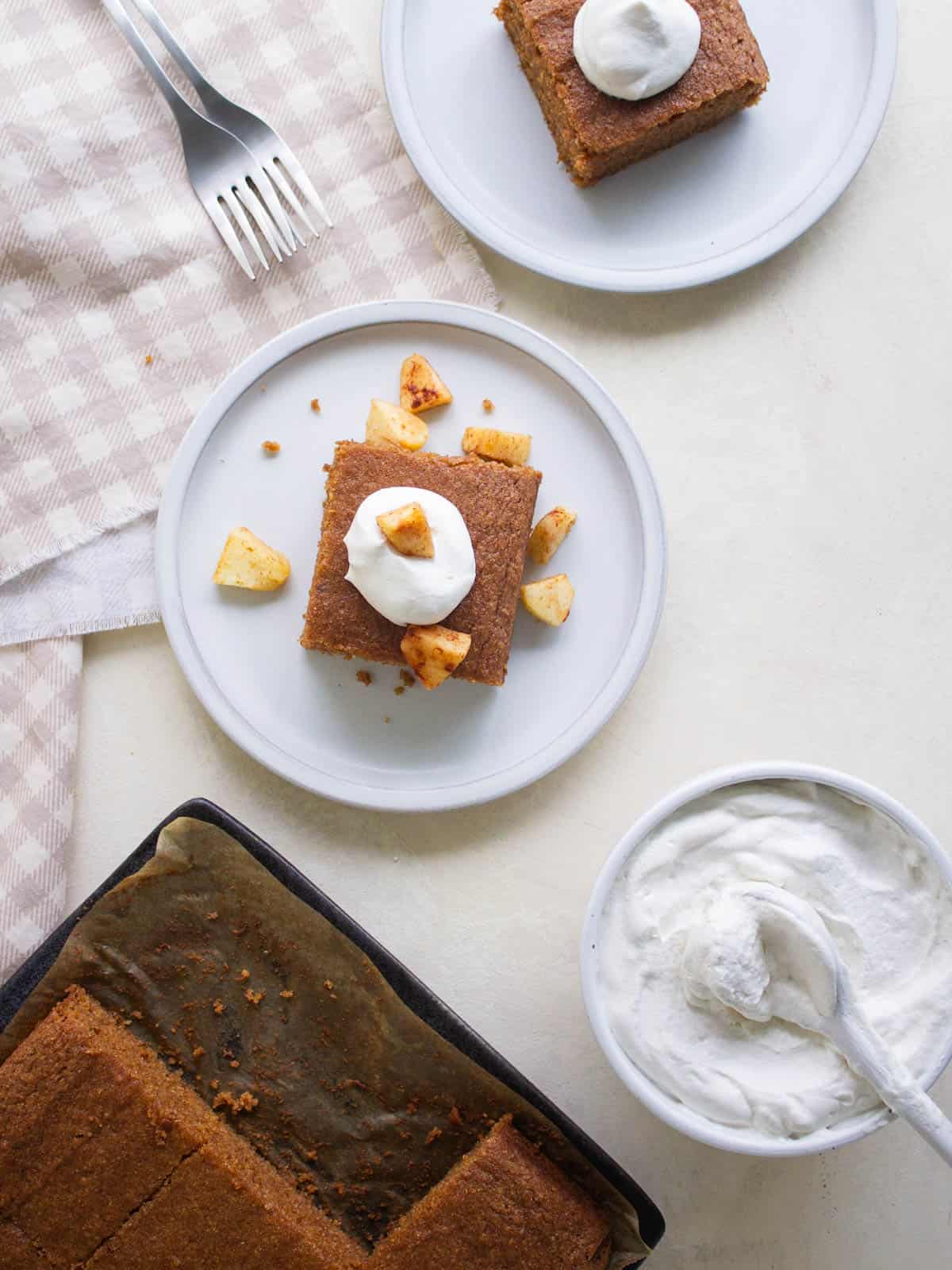 A slice of applesauce cake on a plate with whipped cream and cinnamon apples.