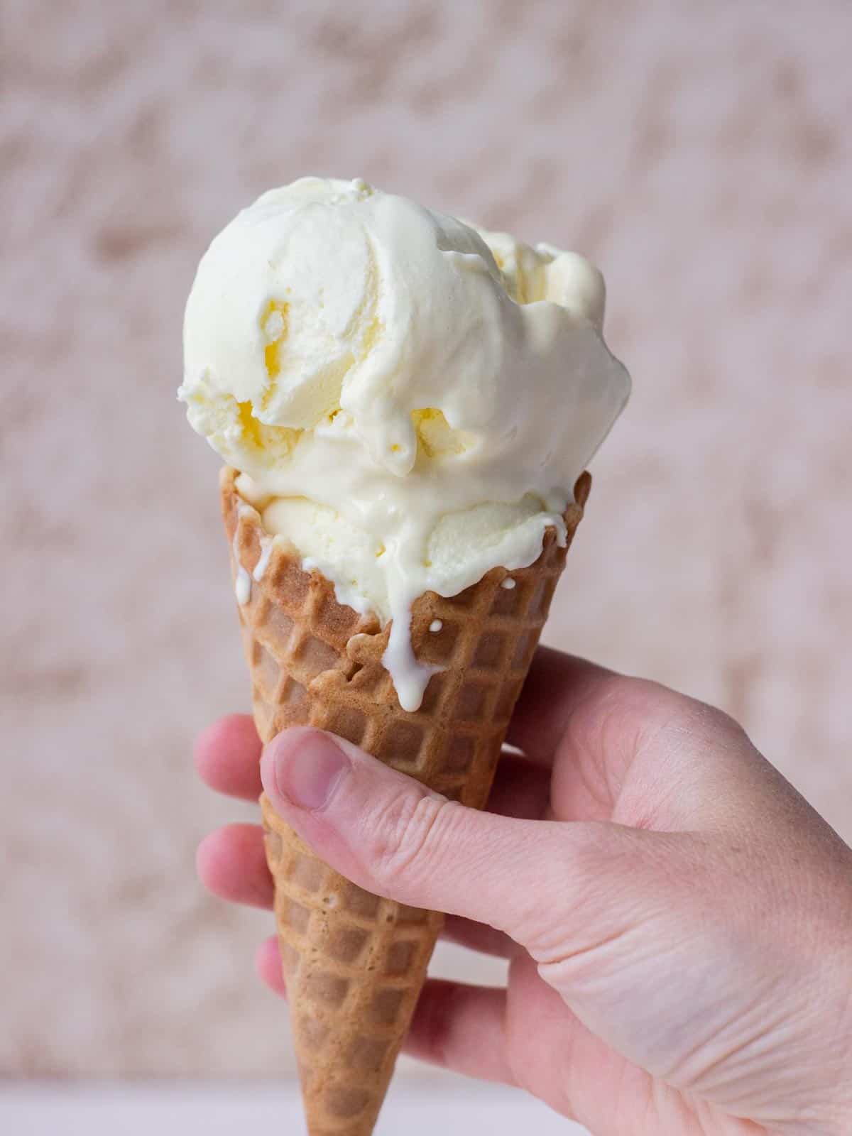 A hand holding a waffle cone with two scoops of vanilla ice cream.