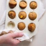 A hand holding a muffin tin full of peach muffins with a towel.