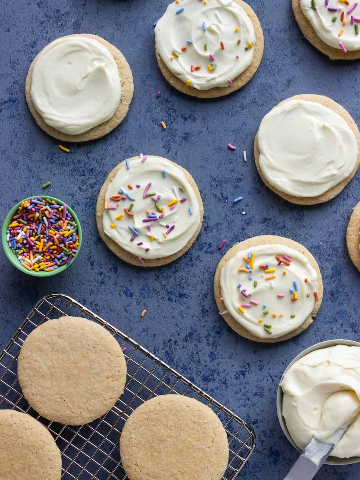 Round sugar cookies arranged randomly some on a cooling rack, some frosted and some frosted with sprinkles.