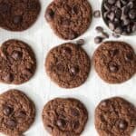 Whole wheat chocolate cookies on a piece of parchment with a bowl of spilled chocolate chips.