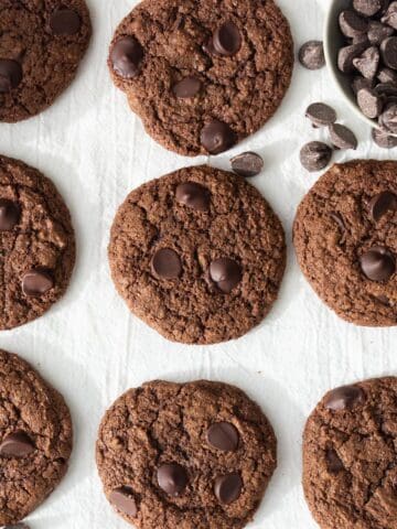 Whole wheat chocolate cookies on a piece of parchment with a bowl of spilled chocolate chips.