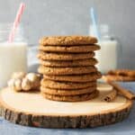 A tall stack of whole wheat molasses cookies on a rustic wooden board with cinnamon sticks, ginger and glasses of milk in the scene.