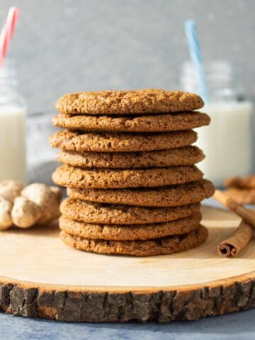 A tall stack of whole wheat molasses cookies on a rustic wooden board with cinnamon sticks, ginger and glasses of milk in the scene.